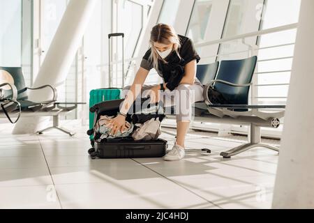 Ritratto di donna con T-shirt nera, maschera bianca monouso, seduta su sedia, vestiario di controllo in valigia nera. Foto Stock