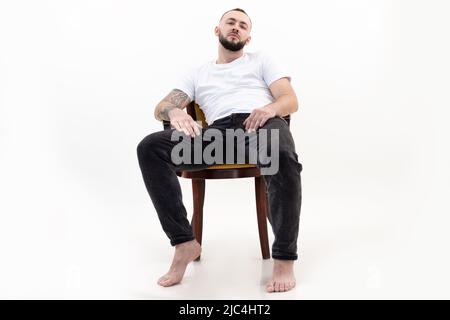 Bruna bella tatuata, uomo a piedi nudi fiducioso e bearded seduta rilassante come capo su sedia come boss. Vista ad angolo basso Foto Stock