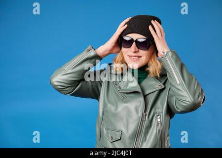 Ragazza gioiosa con cappuccio nero tiene le mani in testa, ha un sorriso divertente, indossando una giacca verde Foto Stock