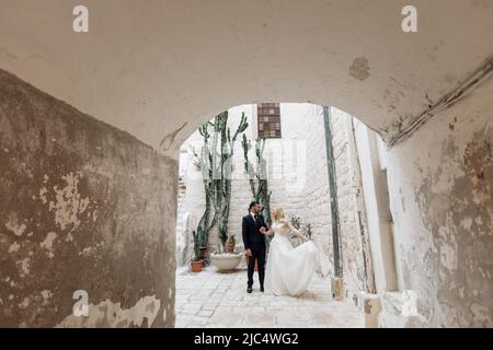 Ritratto all'interno di arco in cemento di giovane sposa bionda in abito bianco e brunet sposo in vestito posa vicino al vecchio edificio della città in Italia, sfondo urbano Foto Stock