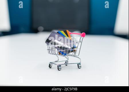 Un mini carrello per lo shopping pieno di carte di credito si trova su un tavolo bianco. Nessuna gente. Primo piano di un carrello con diverse carte di debito bancarie. Foto Stock