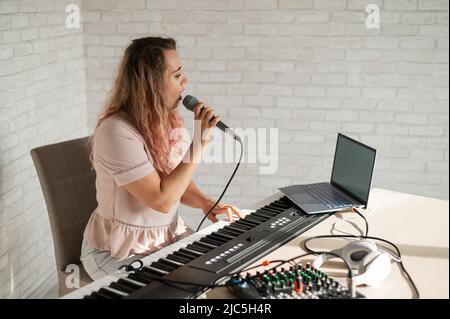 Una donna registra una lezione vocale utilizzando un computer portatile e accompagnando su una tastiera mentre si è a casa. L'insegnante canta una canzone nel microfono e riproduce il Foto Stock