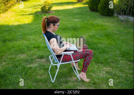 Una donna con occhiali si siede in una sedia pieghevole e si inserisce su un computer portatile. Freelancer femminile lavora a distanza nel giardino del cottage durante la quarantena Foto Stock