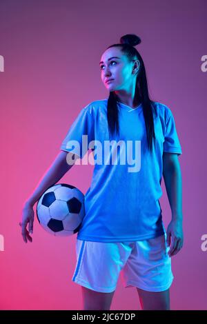 Ritratto di bella ragazza, professionista giocatore di calcio femminile in posa con palla di calcio isolato su sfondo viola studio in luce al neon. Sport Foto Stock