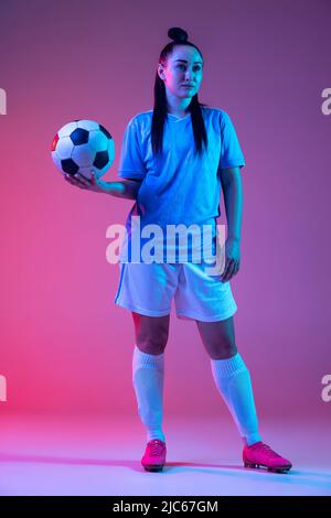 Ritratto di bella ragazza, professionista giocatore di calcio femminile in posa con palla di calcio isolato su sfondo viola studio in luce al neon. Sport Foto Stock