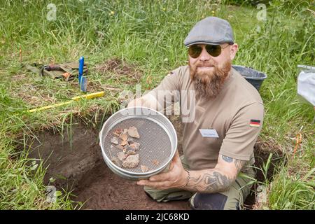 Schenefeld, Germania. 10th giugno 2022. Björn Bigalke, un fabbro e cercatore certificato, si trova in un foro di scavo sul prato della chiesa presso il torrente Meiereibach con un setaccio contenente manufatti di pietra che ha trovato lì. Gli archeologi hanno allestito 20 piccole aree di appena un metro quadrato ciascuna in piazze private e pubbliche per consentire ai residenti di Schenefeld di condurre scavi di ricerca lì sotto guida. Secondo le informazioni, si tratta di un progetto unico in Germania. Credit: Georg Wendt/dpa/Alamy Live News Foto Stock