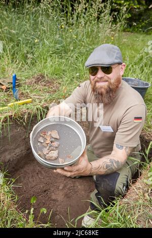 Schenefeld, Germania. 10th giugno 2022. Björn Bigalke, un fabbro e cercatore certificato, siede in un foro di scavo sul prato della chiesa presso il Meiereibach e tiene un setaccio con manufatti di pietra che ha trovato lì. Gli archeologi hanno allestito 20 piccole aree di appena un metro quadrato ciascuna in piazze private e pubbliche per consentire ai residenti di Schenefeld di condurre scavi di ricerca sotto la loro guida. Secondo le informazioni, si tratta di un progetto unico in Germania. Credit: Georg Wendt/dpa/Alamy Live News Foto Stock