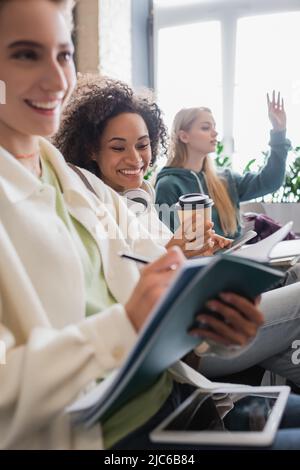allegra donna afroamericana che tiene il caffè per andare vicino blurred la scrittura dell'amico nel taccuino durante la lezione Foto Stock