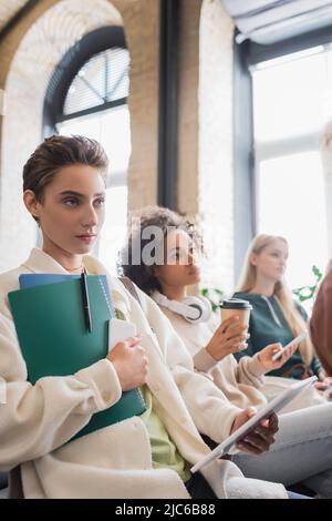 giovane donna con copybooks e gadget ascolto lezione vicino a compagni di classe interrazziali Foto Stock