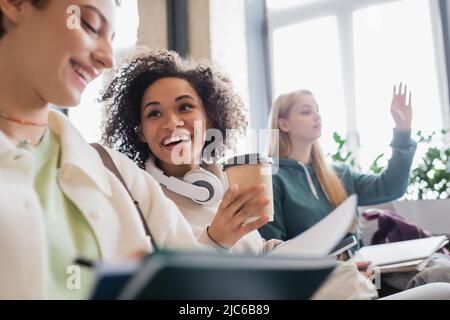 allegra donna afroamericana con cuffie e caffè per parlare con un amico sfocato durante la lezione Foto Stock