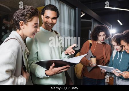 felice uomo afroamericano che mostra copybook a donna vicino amici interrazziali guardando il tablet digitale Foto Stock