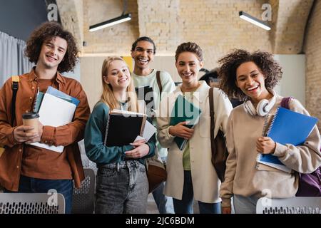 felice studenti multietnici con copybook e gadget che guardano la macchina fotografica in auditorium universitario Foto Stock