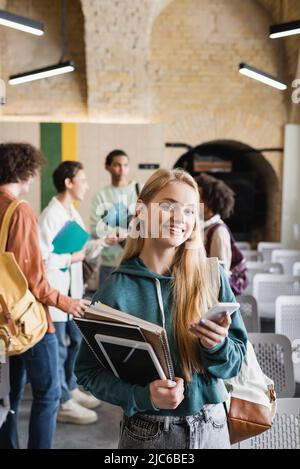 donna felice con notebook, tablet digitale e smartphone vicino sfocato amici multietnici in auditorium Foto Stock