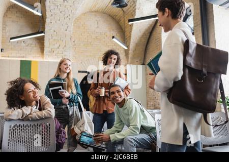 allegri compagni di classe interrazziali con gadget e notebook che guardano lo studente con zaino in pelle in primo piano sfocato Foto Stock