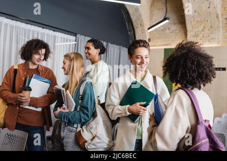 allegri studenti multietnici con copybook e gadget che parlano all'università Foto Stock