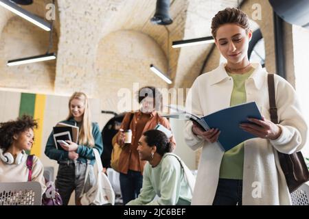 giovane donna che guarda in copybook vicino a studenti multietnici con gadget su sfondo sfocato Foto Stock