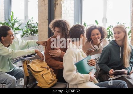 allegri studenti multiculturali con copybook e gadget che parlano mentre si siedono in auditorium Foto Stock