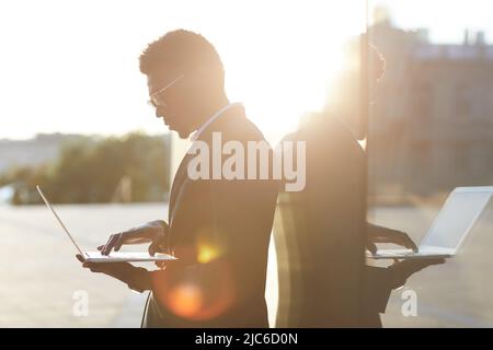 Vista laterale di un giovane uomo serio in vestito, in piedi da un edificio contemporaneo e utilizzando un computer portatile Foto Stock