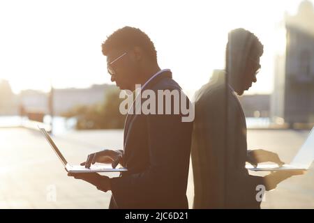 Il giovane uomo d'affari afro-americano attento in occhiali si è concentrato sul progetto in linea che si alza all'aperto e lavora con il laptop Foto Stock