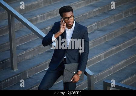 Serio uomo nero mobile in occhiali appoggiati su ringhiera di scala e chiamate sul telefono cellulare in attesa di un collega Foto Stock