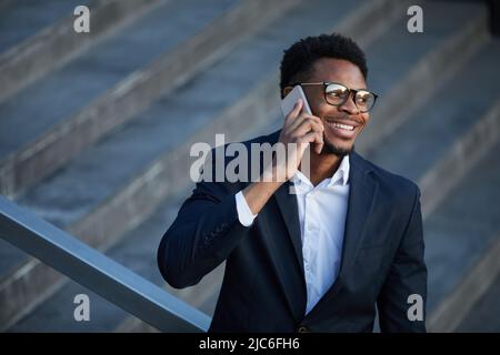 Sorridendo giovane avvocato afro-americano di successo in giacca in piedi sulle scale all'aperto e chiacchierando con il cellulare Foto Stock