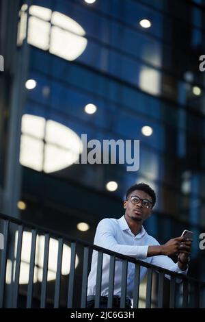 Contemplativo giovane bearded uomo d'affari nero in camicia bianca in piedi sul balcone e usando il gadget, ha lavorato tardi in azienda d'affari Foto Stock