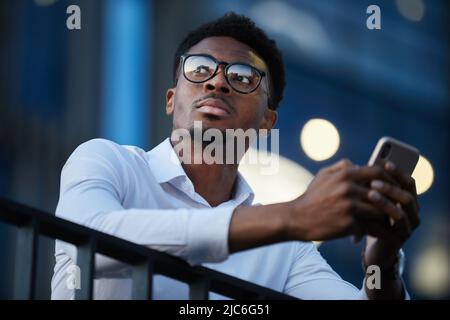 Primo piano del pensivo giovane imprenditore afroamericano con barba in piedi sul balcone e messaggio di testo al telefono Foto Stock