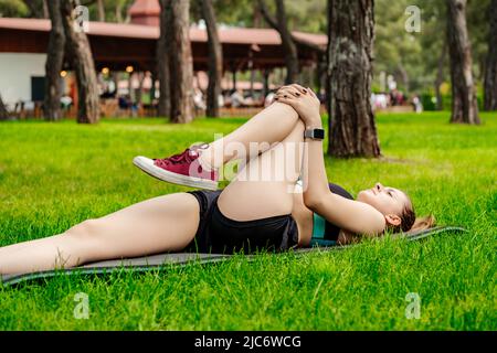 Donna bruna carina che indossa un reggiseno sportivo in piedi sul parco cittadino, all'aperto. Sdraiati in ginocchia per posare al petto, facendo esercizio Apanasana. Concetto di sport all'aperto Foto Stock