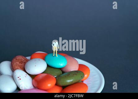 Personaggio in miniatura su fondo grigio isolato di cioccolato dragee. Messa a fuoco selettiva. Essere in cima non è facile, ma alla fine il piacere è sw Foto Stock