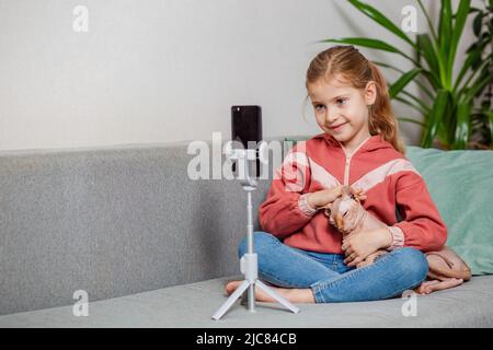 Piccola bionda blogger ragazza, una persona influente, comunica con gli abbonati, conduce una trasmissione dal vivo con un gatto, guarda lo schermo dello smartphone Foto Stock