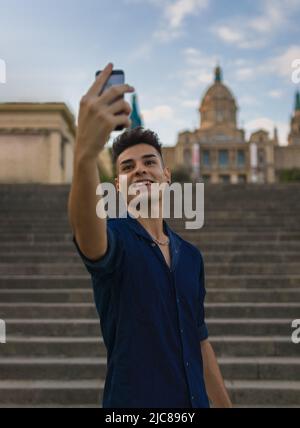 Giovane uomo in piedi sulle scale prendendo un selfie sulla strada con edifici suggestivi e il cielo dietro di lui. Foto Stock