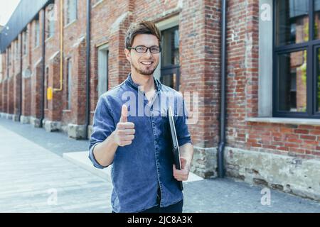 Ritratto di un giovane studente bello, freelance con occhiali. Cammina per le strade della città, tiene un computer portatile nelle sue mani, punta un dito super, l Foto Stock