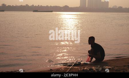 Uomo accovacciato a Riverbank Sunset Vietnam Foto Stock