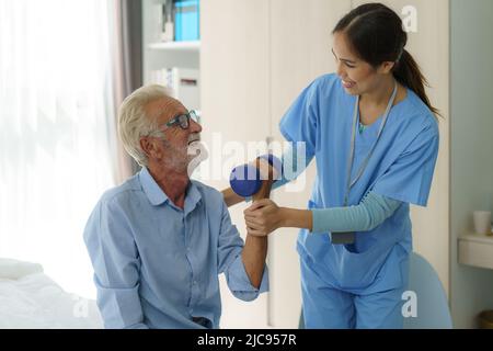Gli esercizi di fisioterapia dell'uomo anziano con la donna asiatica caregiver o fisioterapista dell'infermiera nell'aiuto uniforme del paziente maschio anziano nel paese. Foto Stock
