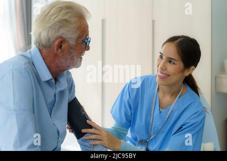 Asian attento operatore sanitario utilizzando il tonometro mentre l'anziano ha problemi di respiro. Medico femminile che misura la pressione sanguigna dell'uomo anziano a casa. Foto Stock