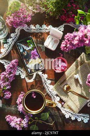 Stile di vita estetico piatto, cuffie senza fili, schermo di telefono, fiori lilla decorazioni e tazza di caffè. Ascoltare musica o audiolibro, prendere Foto Stock