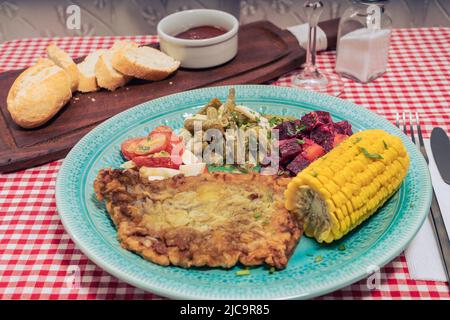 Piatto con schnitzel di carne, insalata di pomodoro, fagioli verdi, barbabietole e uova sode e una pannocchia di mais su un tradizionale tavolo della locanda o del ristorante. Orizzontale Foto Stock