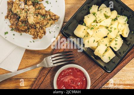 Deliziosa insalata di patate e prezzemolo in una ciotola nera, morsi di manzo e una pentola di salsa Ketchup su un asse di legno. Foto Stock