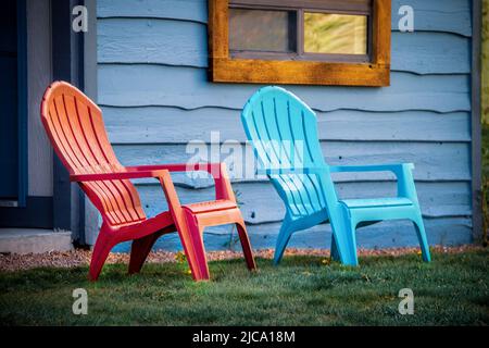 Primo piano di sedie a arondiack rosse e turchesi seduti all'esterno della porta di cabina di legno blu sull'erba. Foto Stock