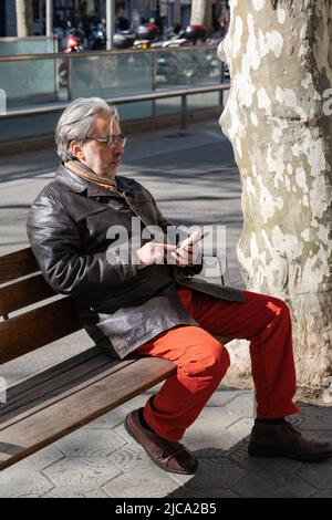 L'uomo maturo con capelli bianchi e barba seduti su una panca di legno utilizza il telefono. Relax in un ambiente urbano. Foto Stock