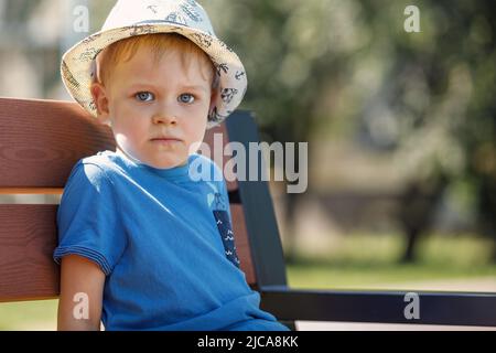 Ritratto di un ragazzo carino con un cappello bianco e una camicia blu. Un bambino siede su una panca nat nel parco e guarda la telecamera Foto Stock