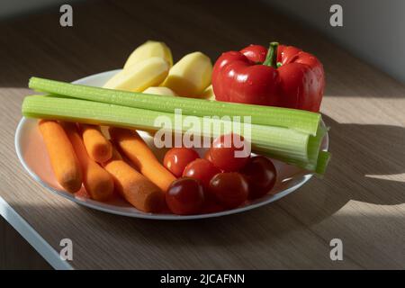 Piatto bianco con pomodori ciliegini, carote, sedano, peperoni e patate alla luce del sole su tavola di legno. Foto Stock