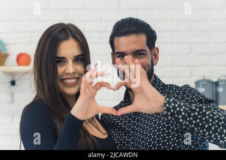 Interrazziale felice bella giovane coppia piegando le mani in una forma di cuore, sorridendo e guardando la macchina fotografica, in piedi in una cucina. Foto di alta qualità Foto Stock