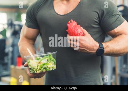 Muscoloso irriconoscibile vestito ragazzo caucasico in una t-shirt che tiene una ciotola di insalata di verdure verdi e un falso cuore umano. Foto di alta qualità Foto Stock