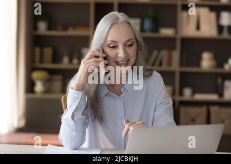 La donna d'affari anziana attrattiva fa chiamata di affari seduta al posto di lavoro Foto Stock