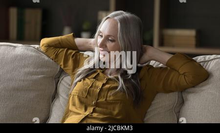 Donna di mezza età con lunghi capelli grigi seduta su un comodo divano Foto Stock