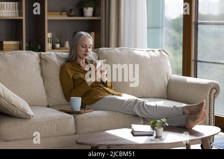 Donna di mezza età che si rilassa sul divano con uno smartphone moderno Foto Stock