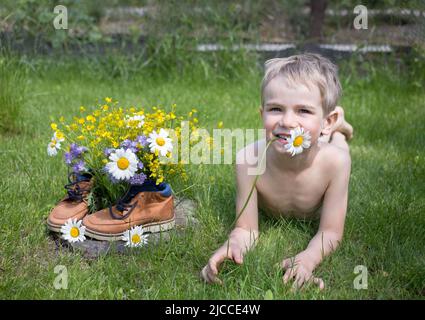 bambino ragazzo si trova sull'erba, accanto ad un bouquet raccolto di fiori selvatici in piedi in stivali. gioia, atmosfera positiva, buona infanzia. Ciao estate. S Foto Stock