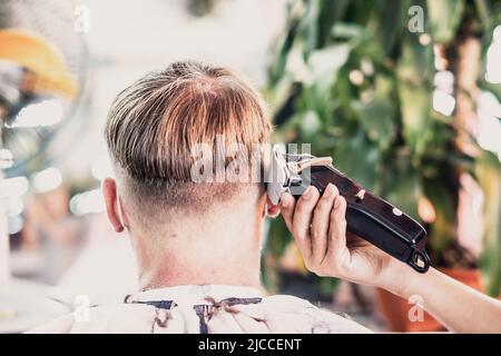 Salone fotografico Lifestyle. Il cliente giovane indossa la maschera facciale, vista posteriore laterale. Regolacapelli elettrico per pettini da lavoro maschio professionale Foto Stock