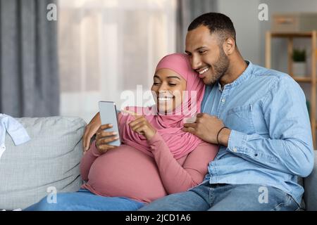 Felice coppia islamica nera incinta usando lo smartphone mentre si rilassa insieme a casa Foto Stock